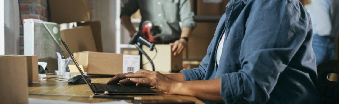 Business owner typing on laptop in warehouse packing boxes